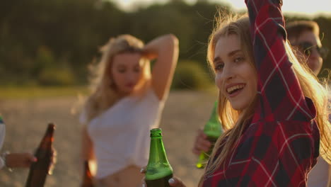 A-young-female-is-dancing-on-the-open-air-party-with-her-friends-on-the-beach.-Her-long-blonde-hair-is-flying-on-the-wind.-She-smiles-and-enjoys-a-summertime-on-the-lake-coast-with-beer.